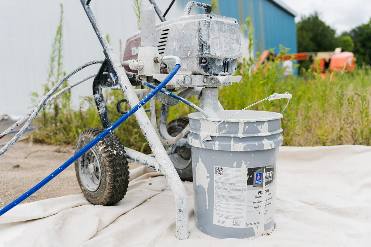 PMV - Blog - Can You Paint Vinyl Siding 2 paint sprayer outside using a large 5 gallon bucket of paint.