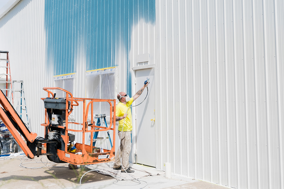 Commercial painter from PMV Painting using a paint sprayer on the exterior of a metal building during an exterior commercial repaint project in Kalamazoo Michigan